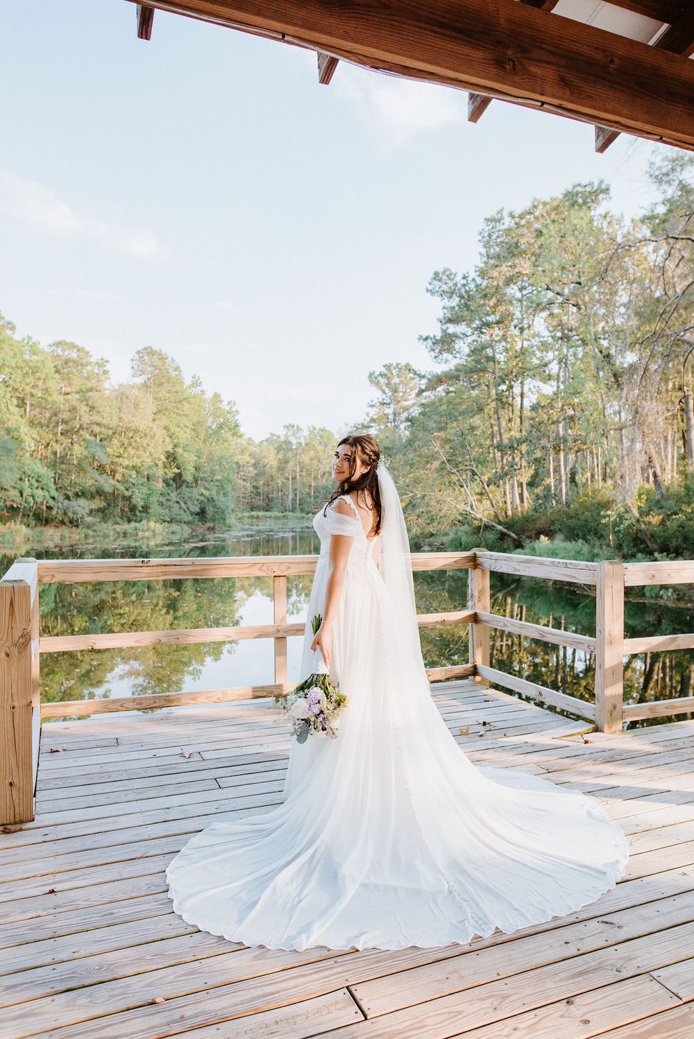 A bride in a flowing white dress with long train stands on a wooden deck overlooking a serene pond surrounded by pine trees.