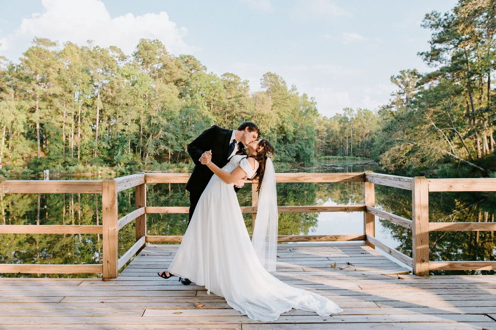 A romantic wedding couple shares a tender moment on a wooden dock overlooking a serene lake during golden hour.