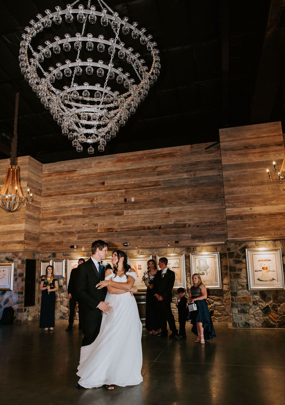 A couple performs their first dance under elegant crystal chandeliers in a rustic wooden reception venue.