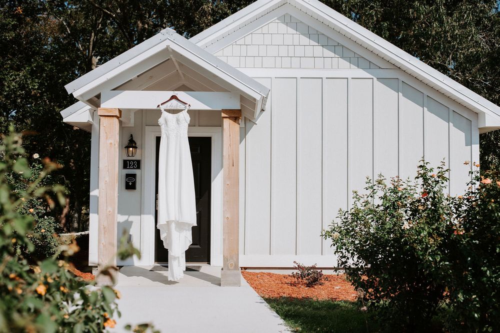 A white wedding dress hangs in the doorway of a charming white cottage-style building with a small front porch.