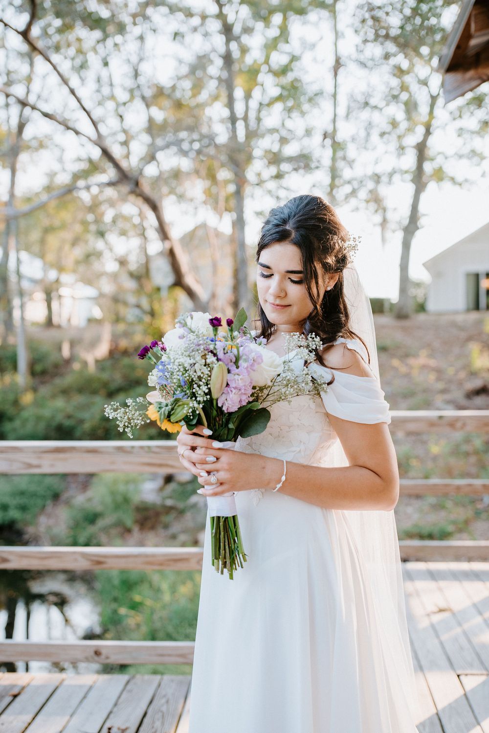A bride in a white off-shoulder wedding dress holds a delicate bouquet of white and purple flowers in an outdoor setting.