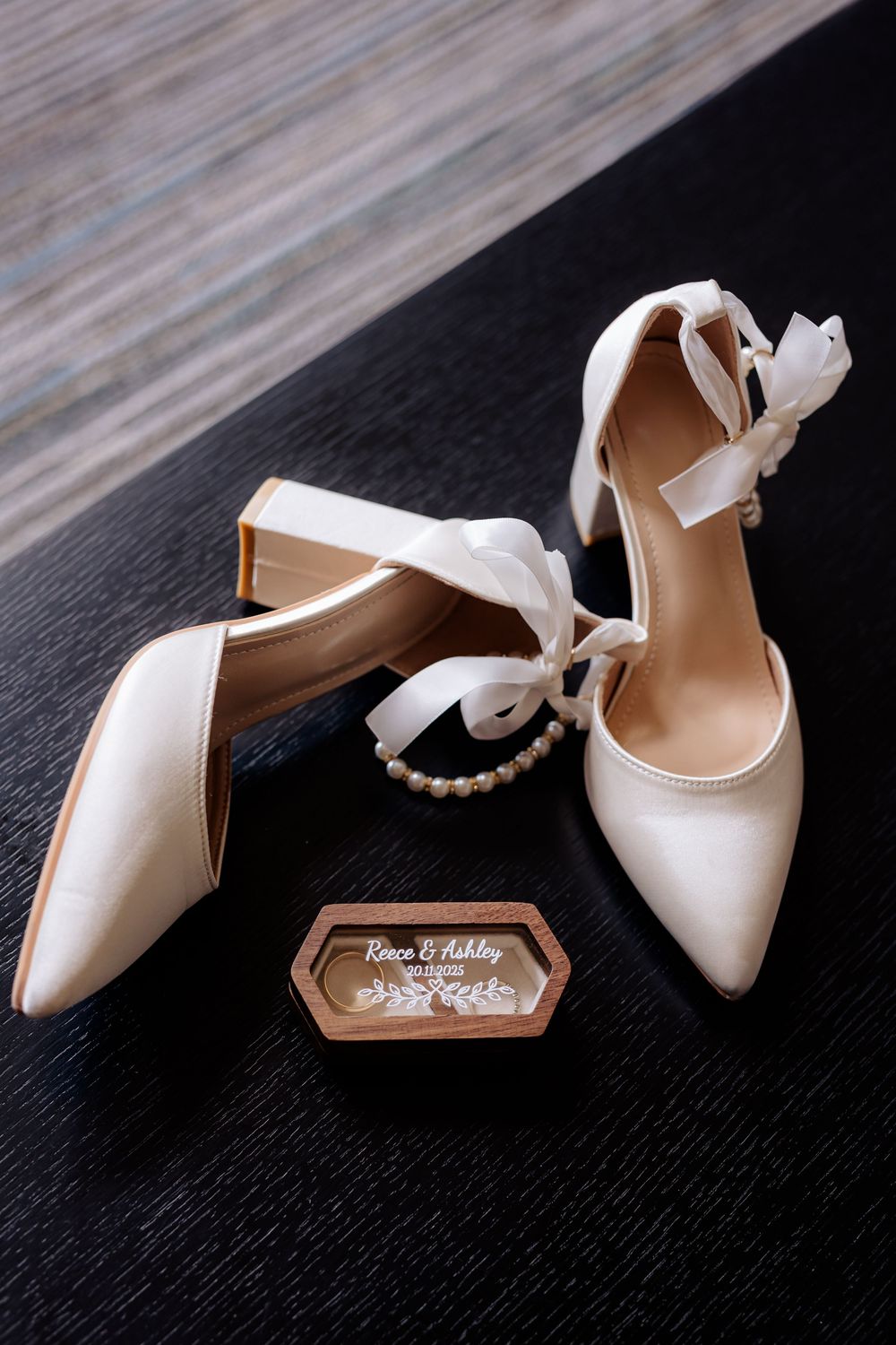 Elegant white bridal shoes with ribbon ties displayed next to a hexagonal ring box on dark surface.
