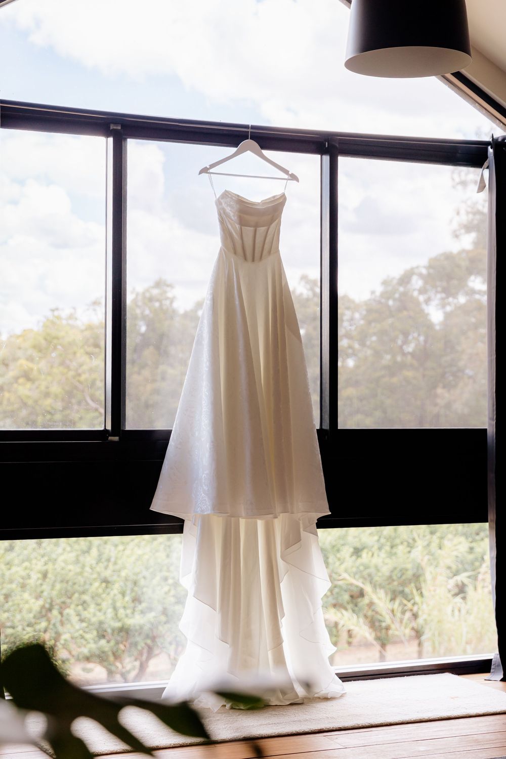 White wedding dress hanging in window creating elegant silhouette against natural light.