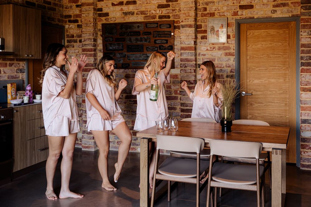 Ballet dancers in white tutus stretch at wooden barre against exposed brick wall during dance class rehearsal.