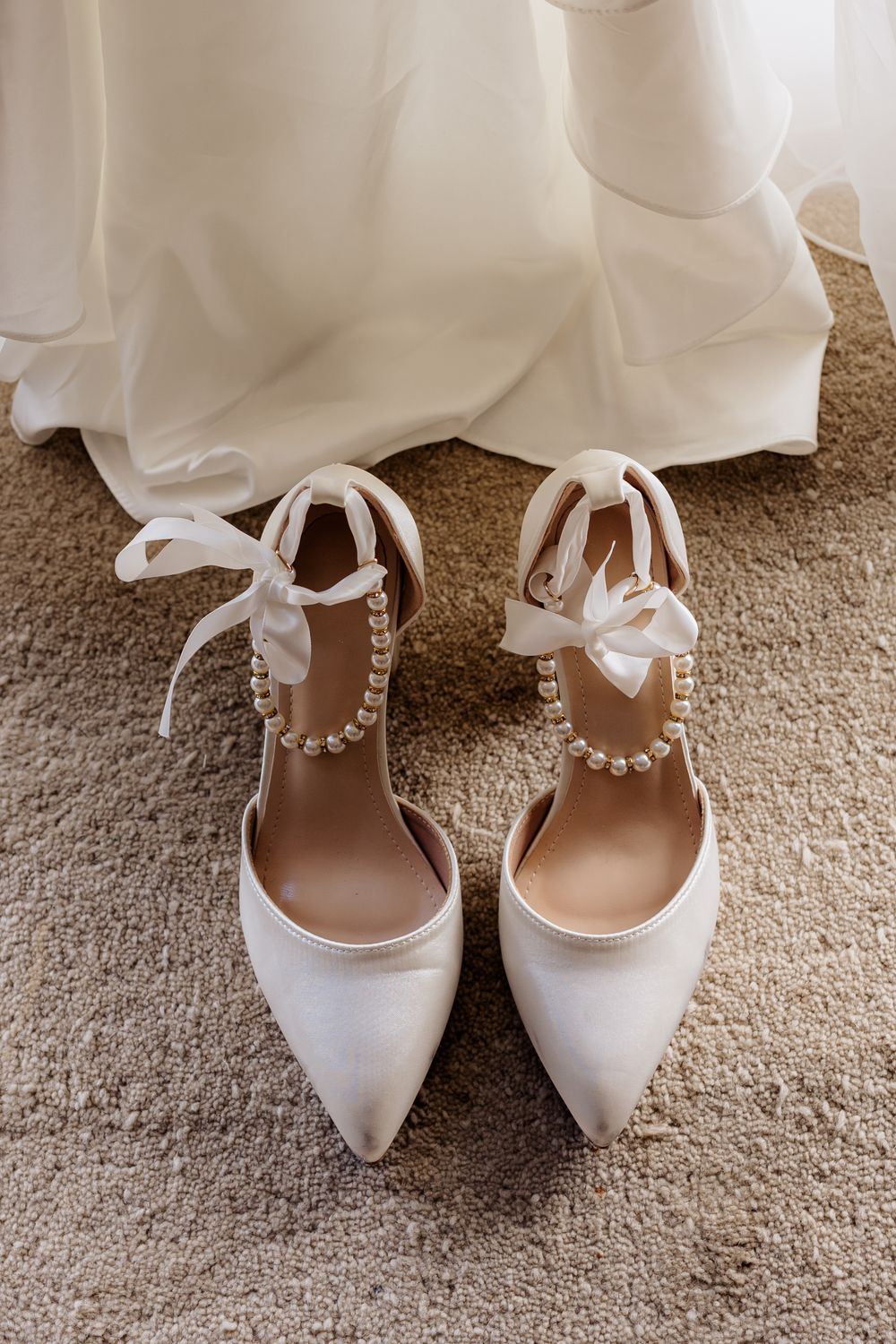 Elegant white bridal shoes with delicate bow details displayed on beige carpet.