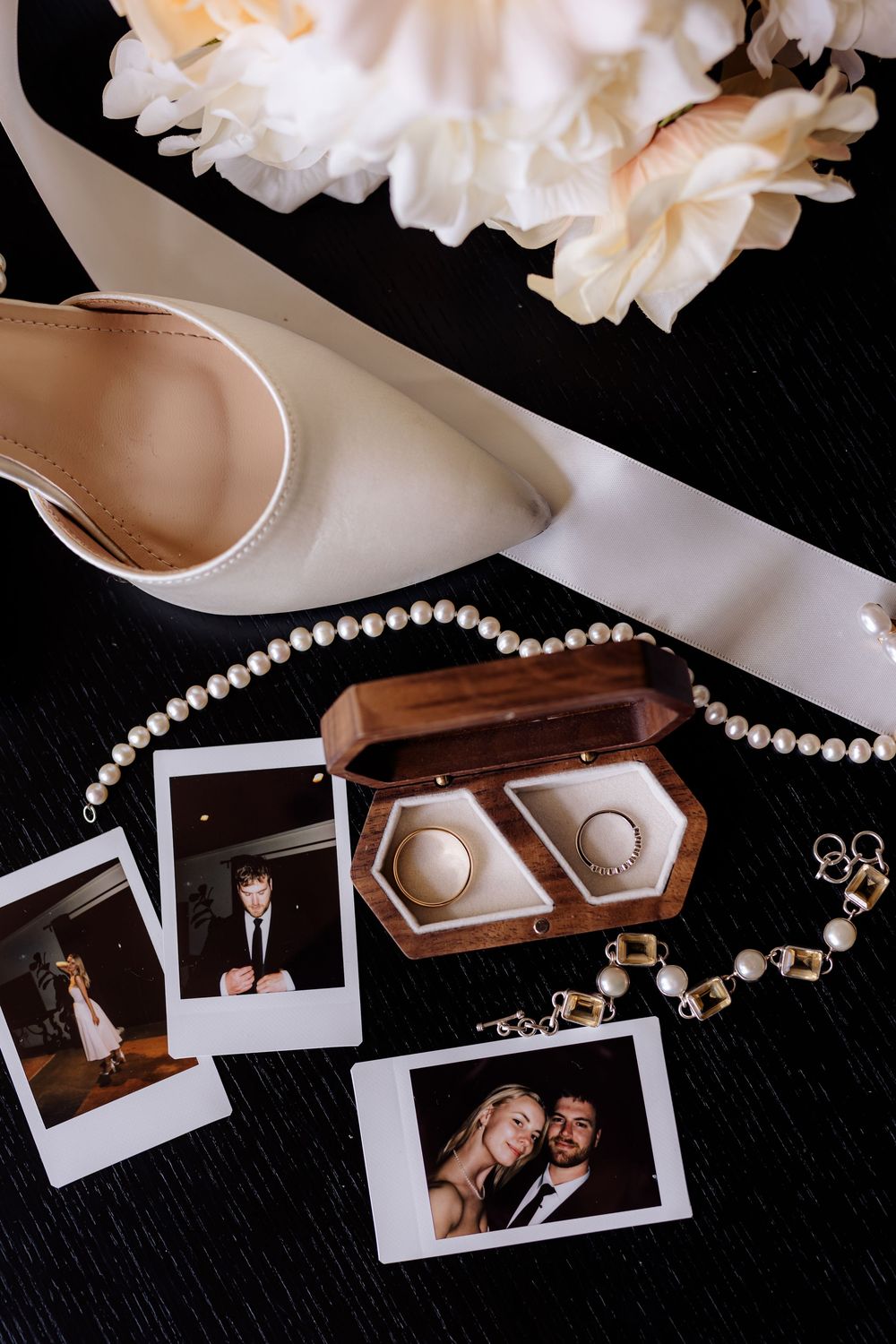 Wedding details including pearl necklace, rings, and polaroid photos arranged on dark background.
