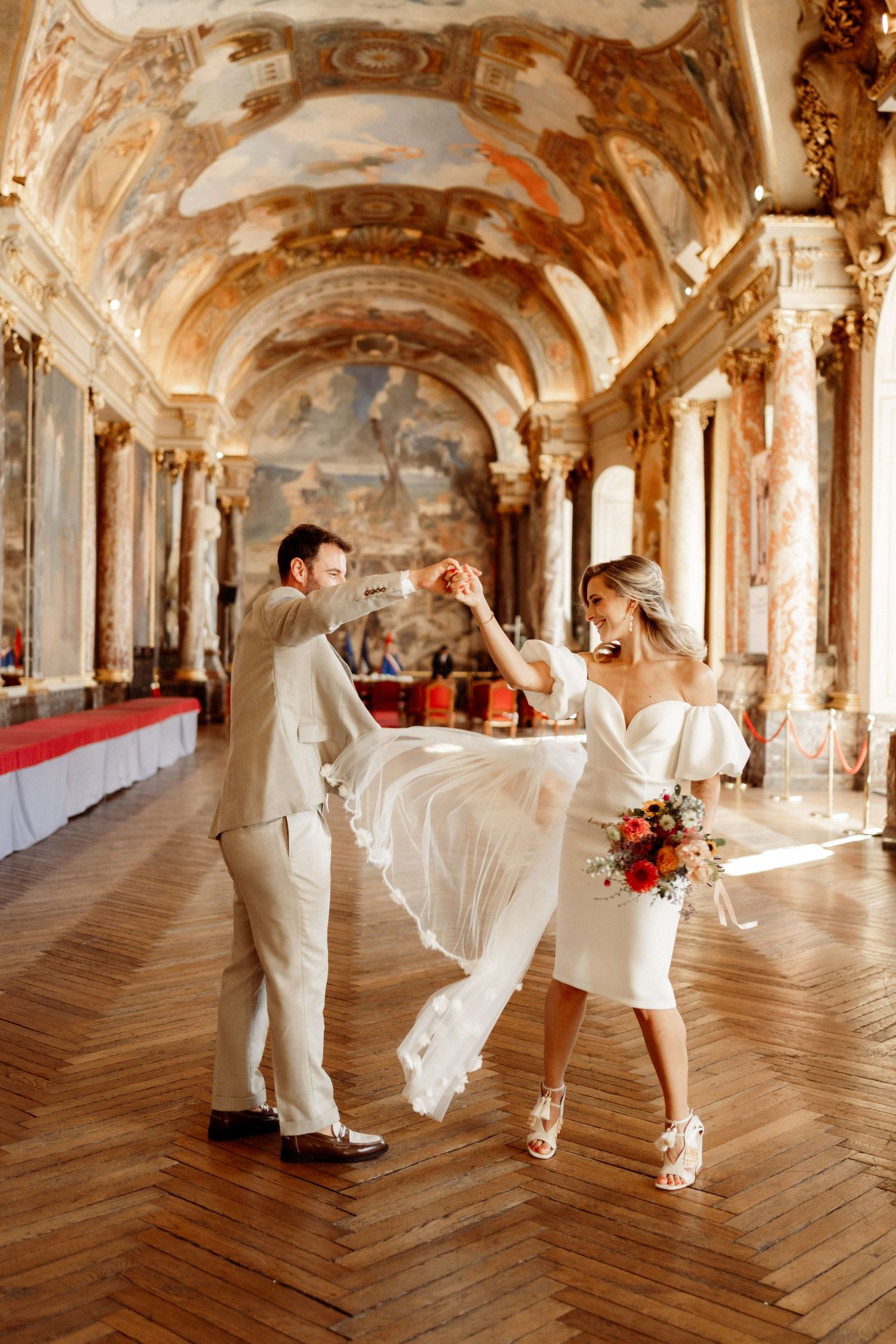 A couple dances in an ornate baroque hall with frescoed ceilings and herringbone floors.