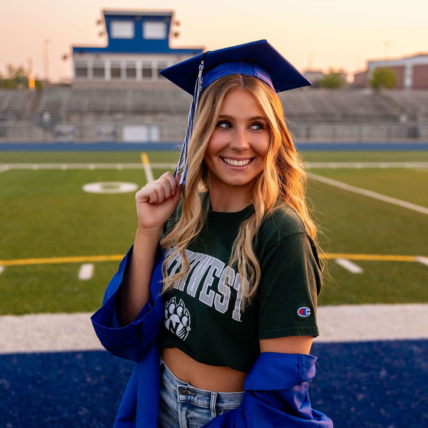 Graduate in blue cap and gown poses on football field during graduation photo session in Bennington, NE. She's wearing her college tshirt.