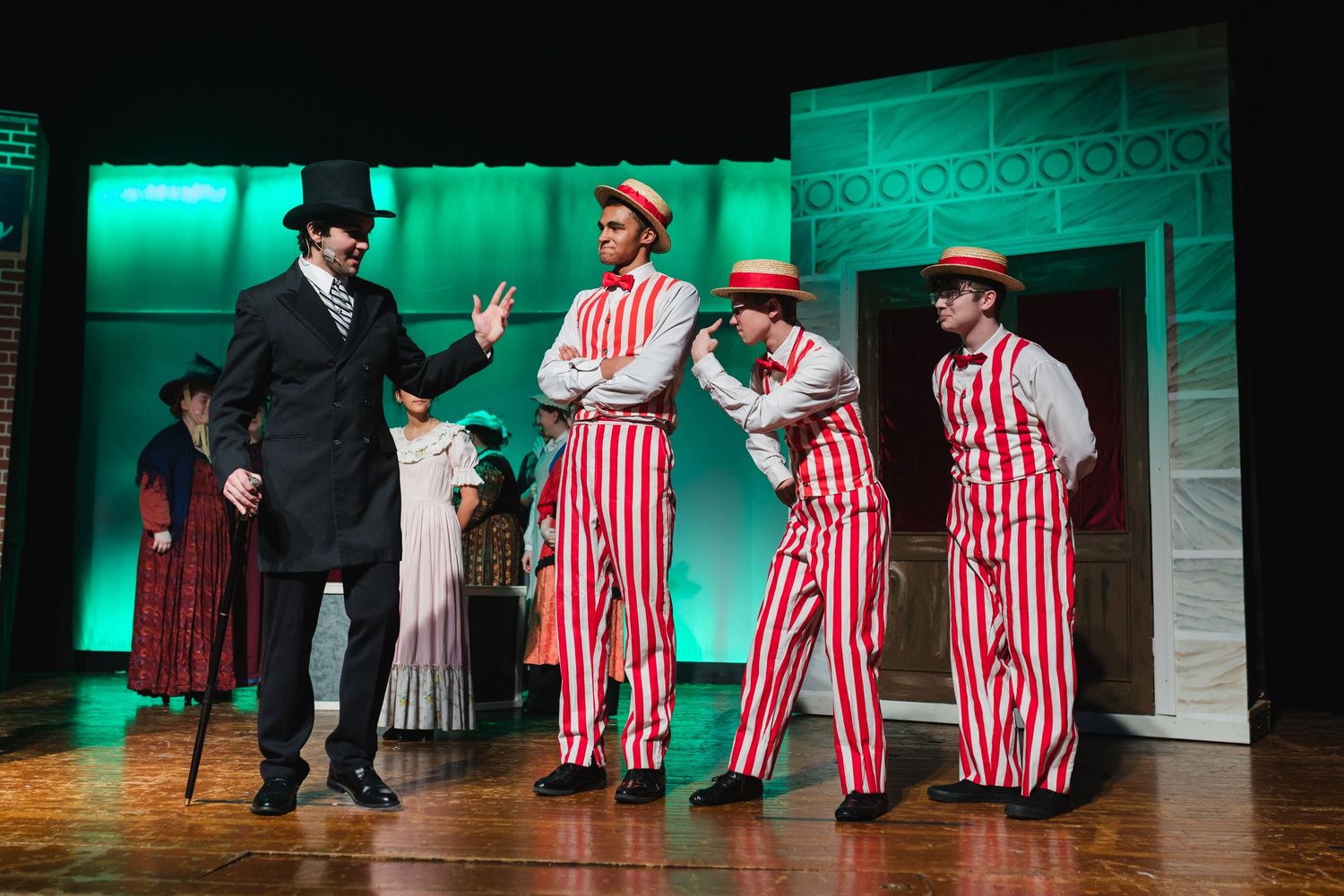 Performers in red and white striped costumes dance and interact on a theater stage with green lighting effects.