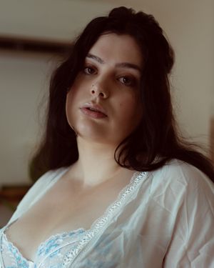 Portrait in soft lighting wearing a white lace-trimmed top with dark hair against a neutral background.