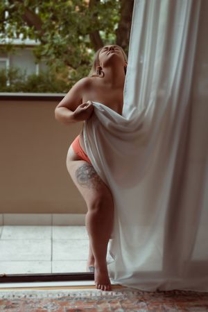 Artistic boudoir photo showing a person wrapped in flowing white curtains against natural window light in an intimate setting.