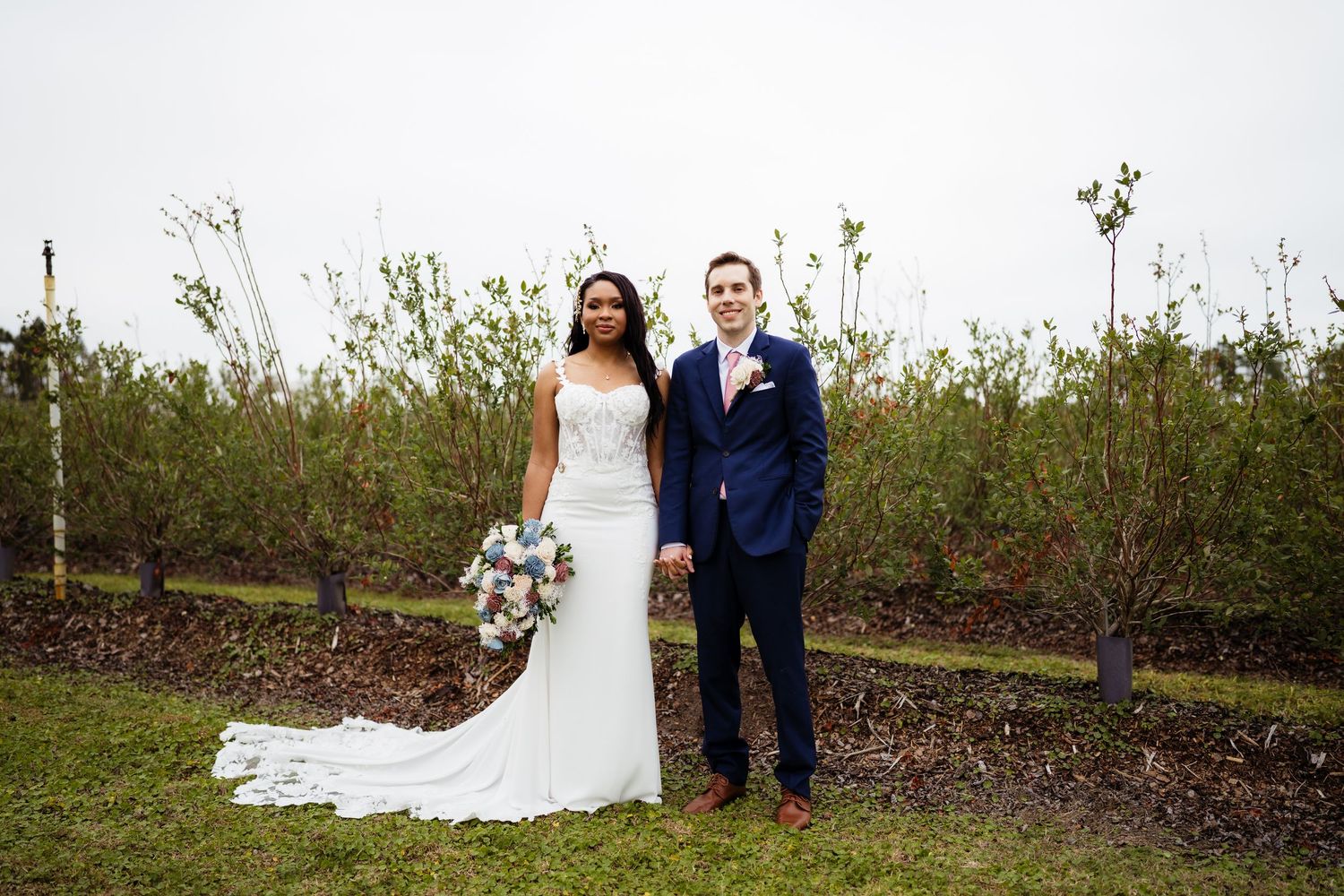 A couple in wedding attire stands together in a natural outdoor setting with greenery and trees in the background.
