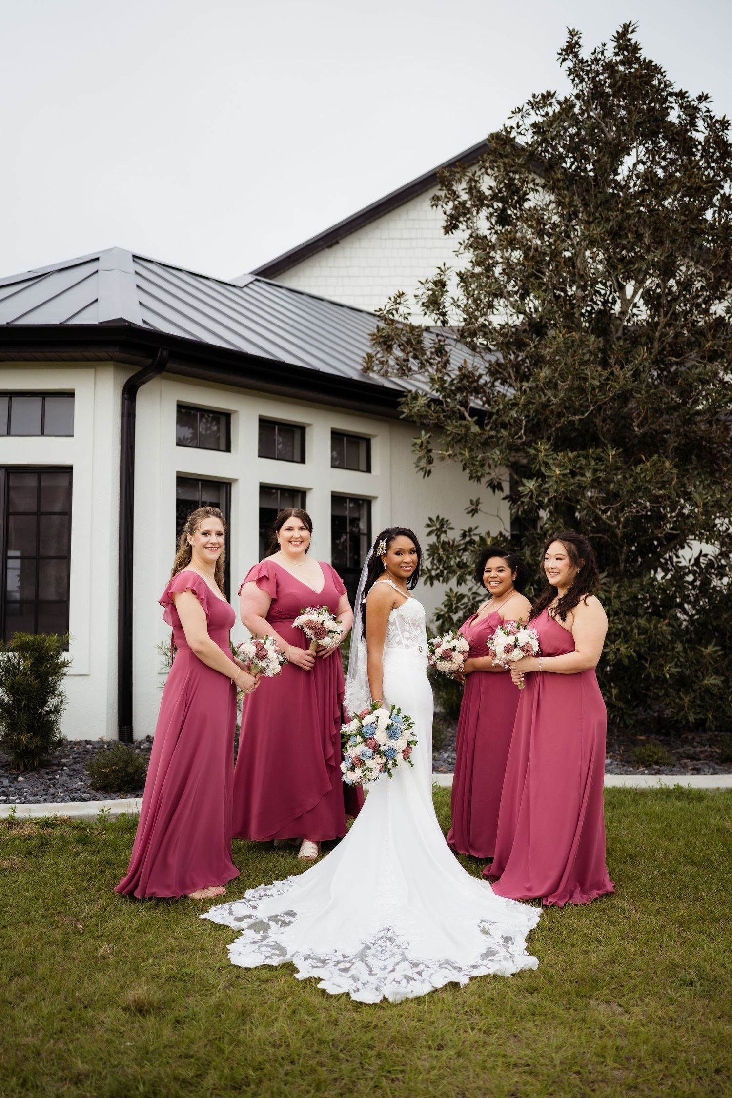 Bride in white gown poses with bridesmaids wearing burgundy dresses on a grassy lawn outside a white building.
