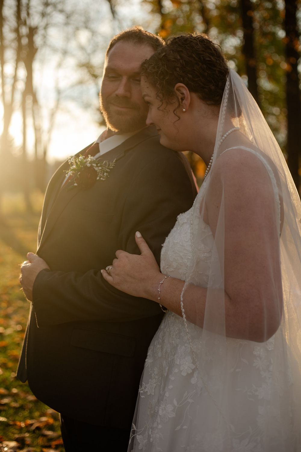 A bride in a white dress and veil stands close to someone in black during a golden sunset among autumn trees.