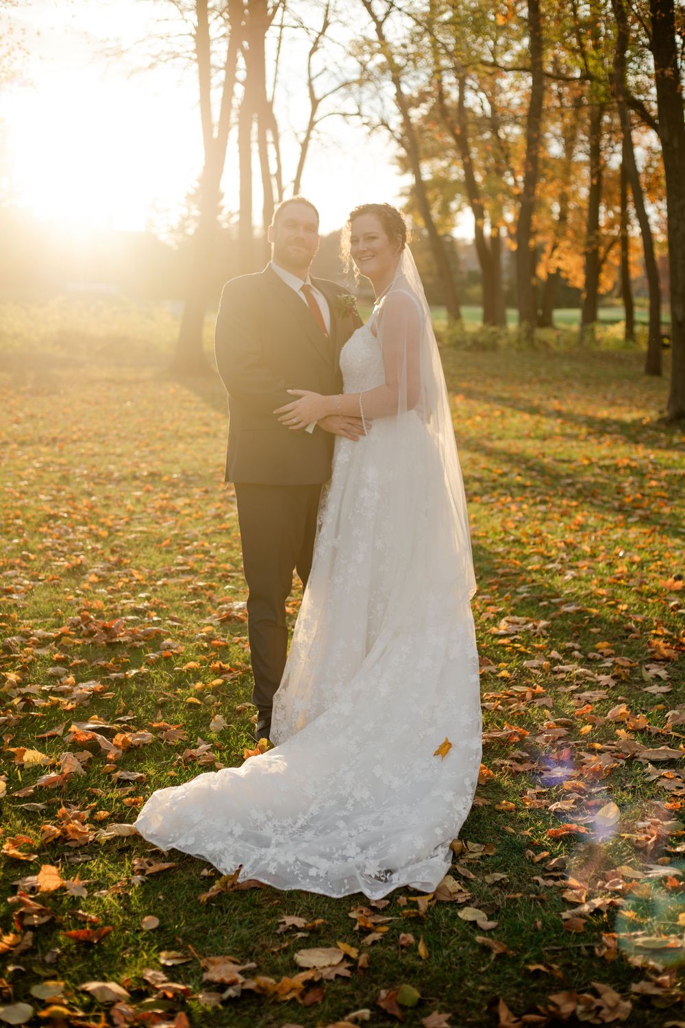 A romantic wedding couple stands among fallen autumn leaves in a sunlit forest during golden hour.