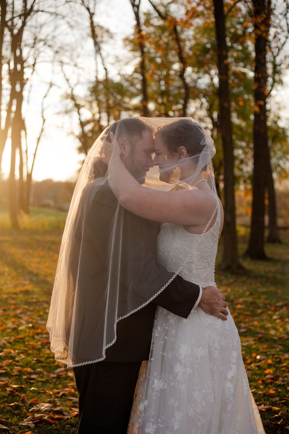 A romantic wedding couple embraces during golden hour in an autumn forest setting with fallen leaves.