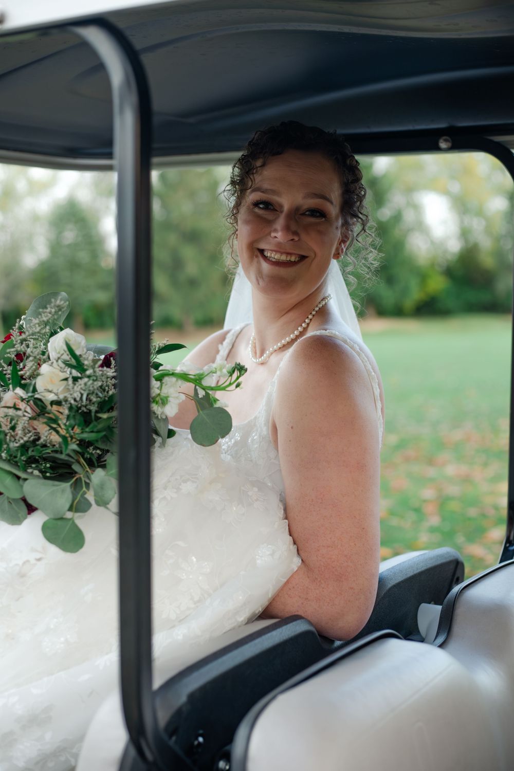 A bride in a white gown sits in a golf cart holding a bouquet with eucalyptus and white flowers.