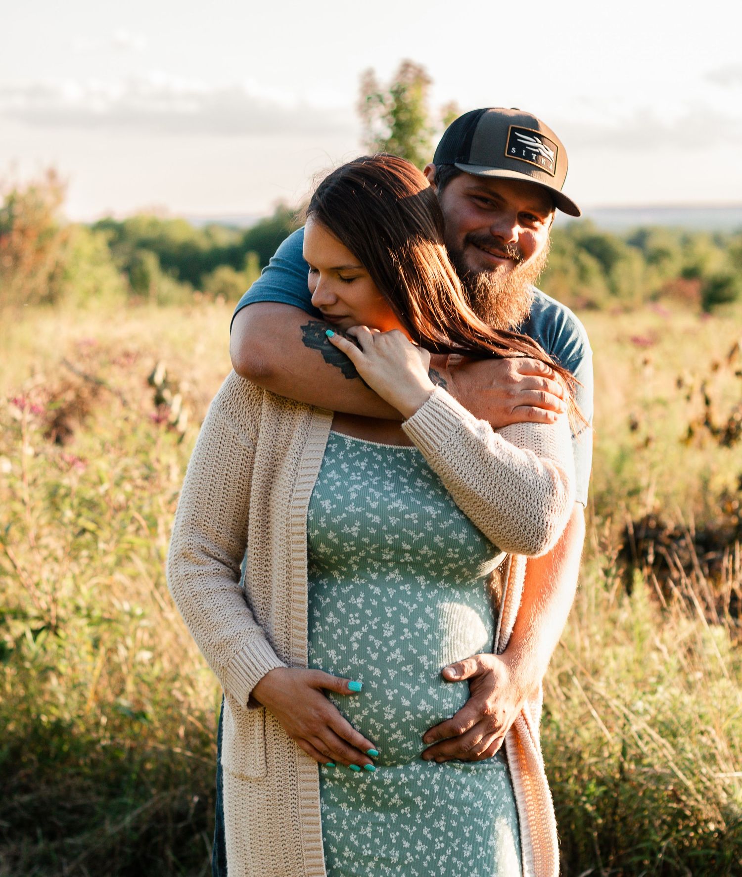A couple embraces in a sunlit meadow during a maternity photoshoot at golden hour.