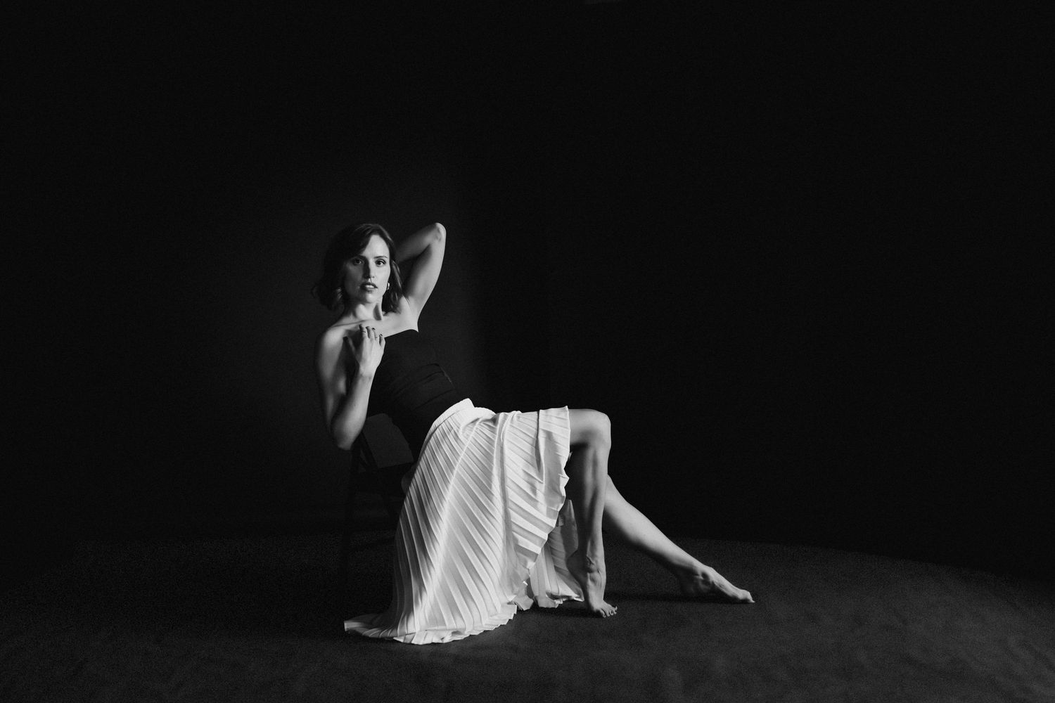 Black and white fine art photo of woman seated on floor in white skirt against dark background.