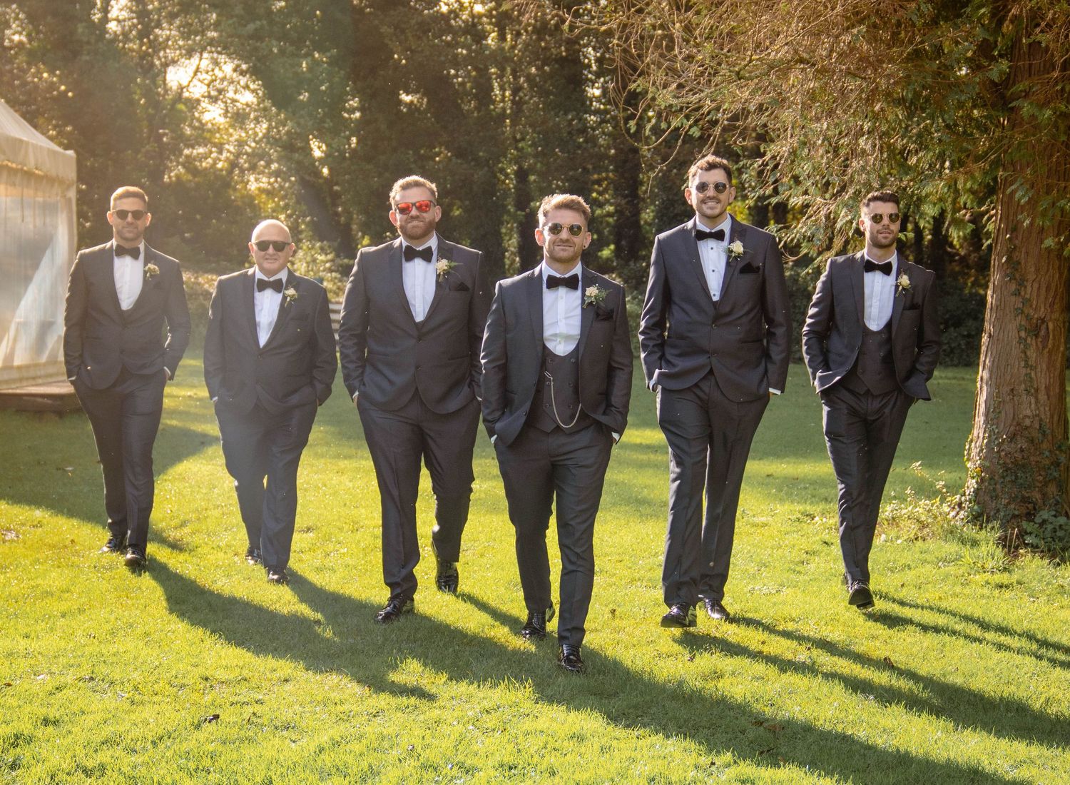 Groomsmen in black tuxedos walk in a line across a sunlit lawn during a wedding celebration.