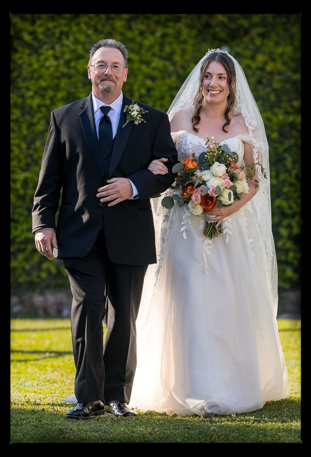 A traditional wedding portrait outdoors with a bride in white dress holding a colorful bouquet alongside a companion in a dark suit.