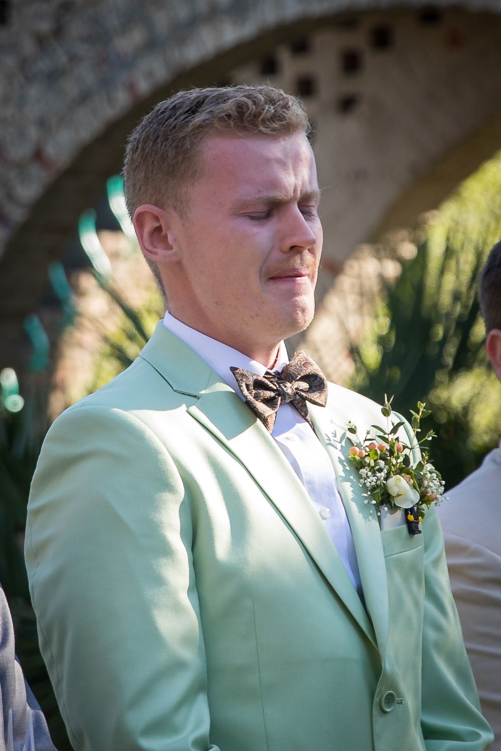 Groomsman in mint green suit jacket and bow tie stands outdoors during a wedding ceremony.