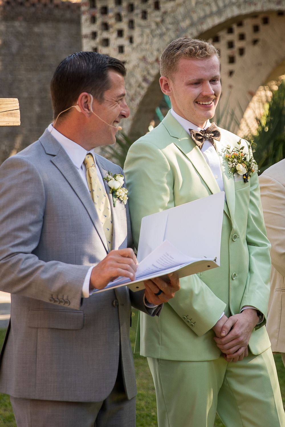 Two men in formal attire, one wearing a gray suit and one in mint green, read from papers during an outdoor ceremony.