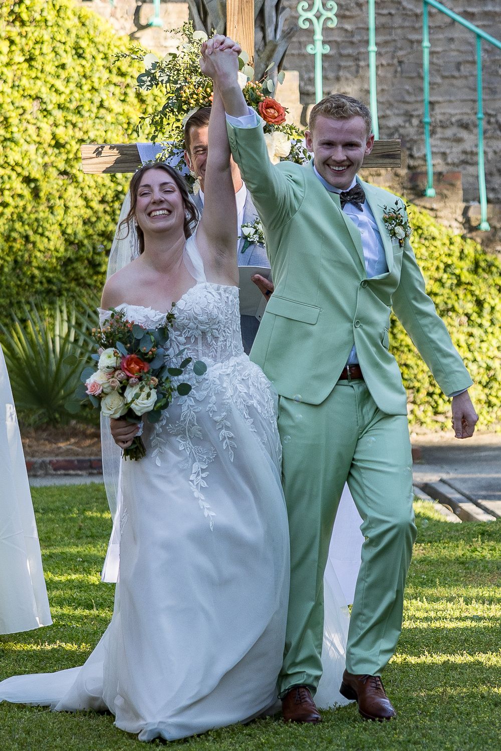 A joyful wedding celebration outdoors with a couple in a white gown and mint green suit raising their hands in triumph.