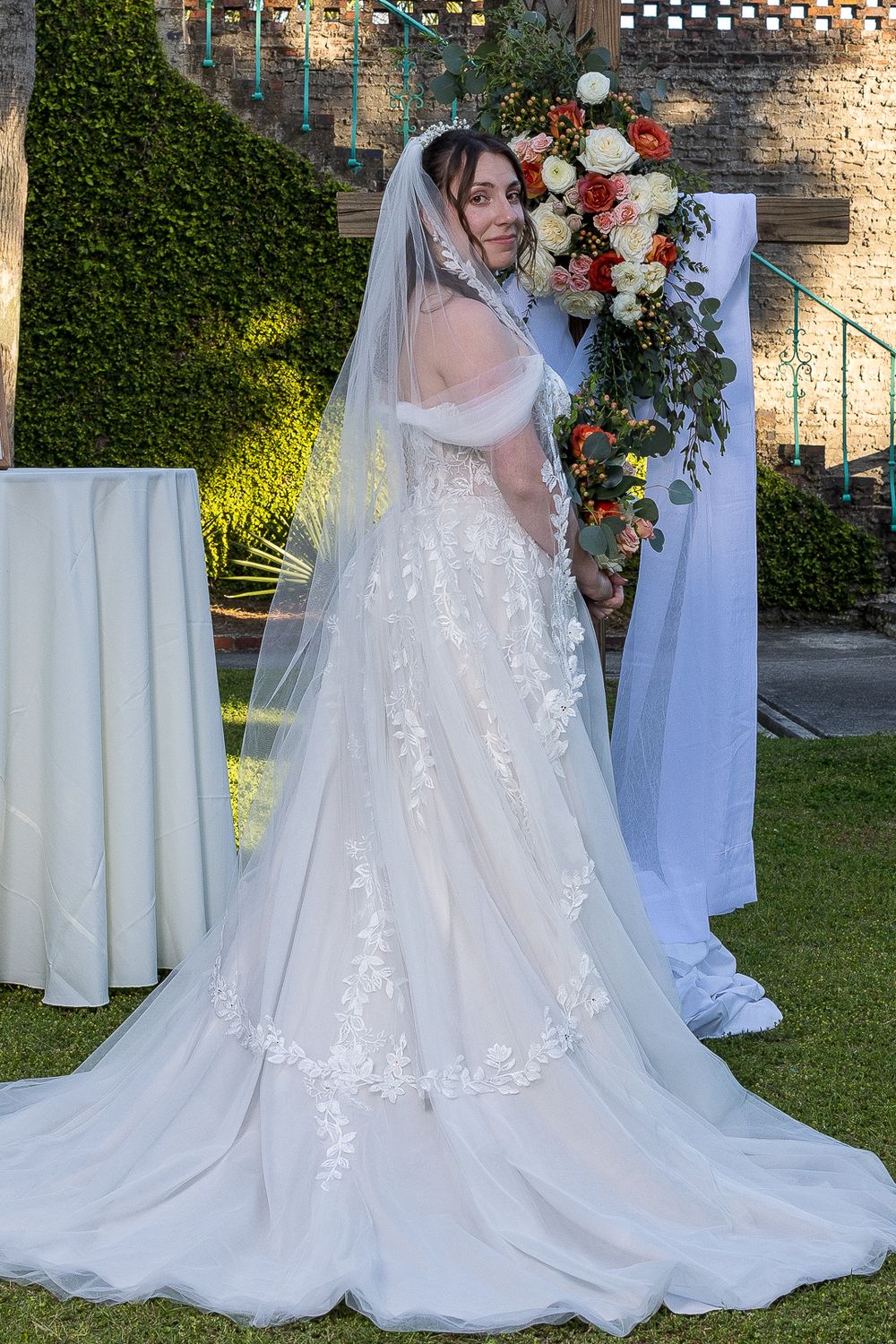 A bride in a flowing lace wedding gown with a long veil poses in front of a floral arch in an outdoor garden setting.