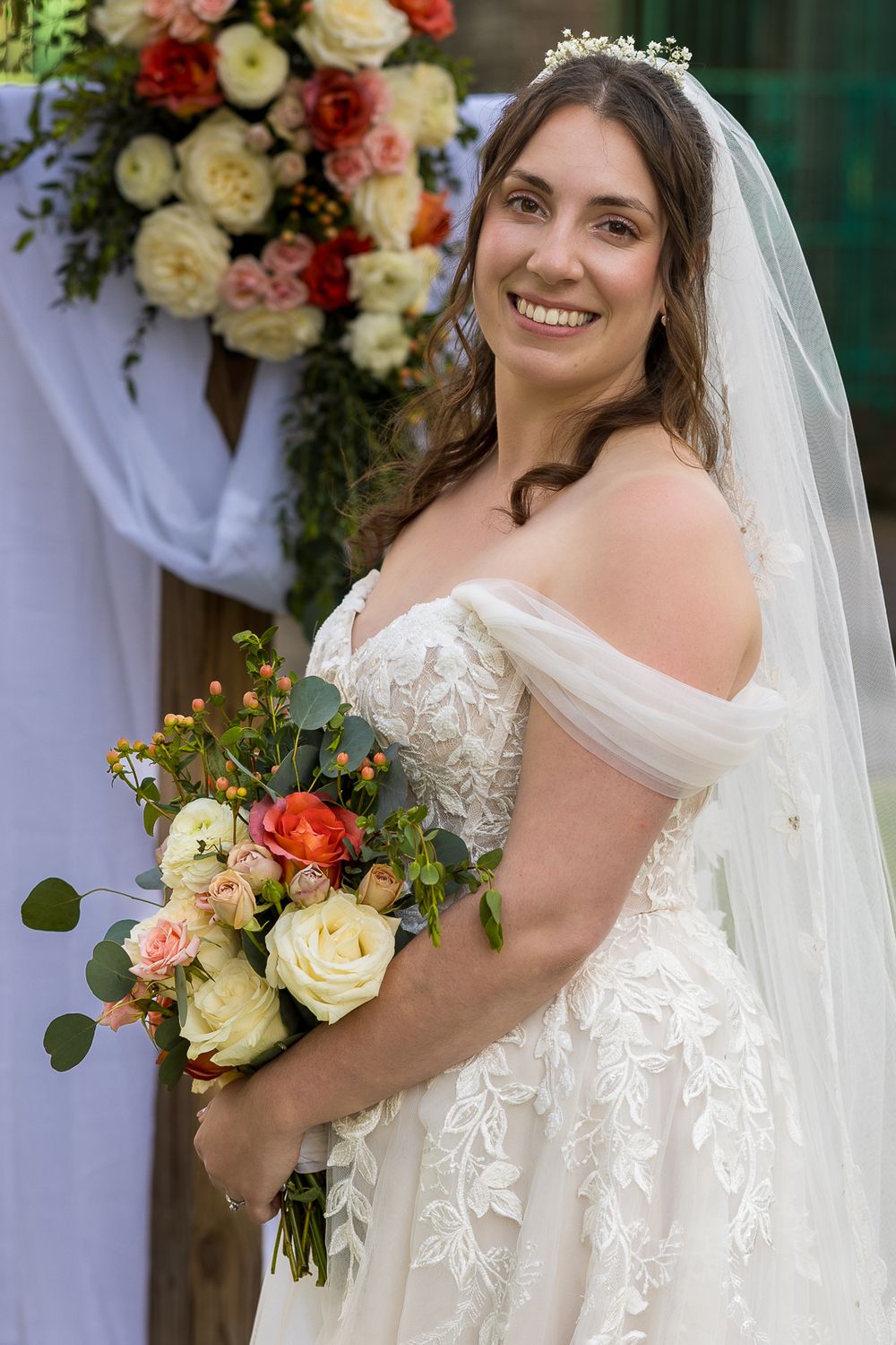 A bride in a lace off-shoulder wedding dress holds a bouquet of pink and cream roses next to floral decorations.