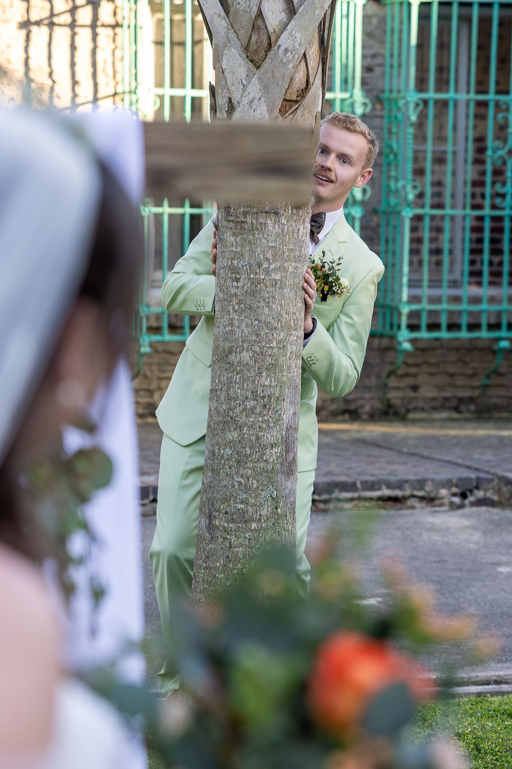 A person in a mint green suit stands near a tree trunk during an outdoor wedding ceremony.