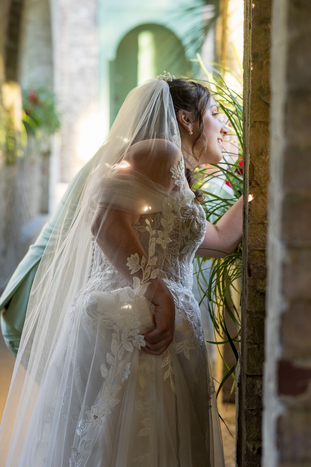 A bride in an elegant white lace gown and veil stands in soft natural light near a stone wall with greenery.