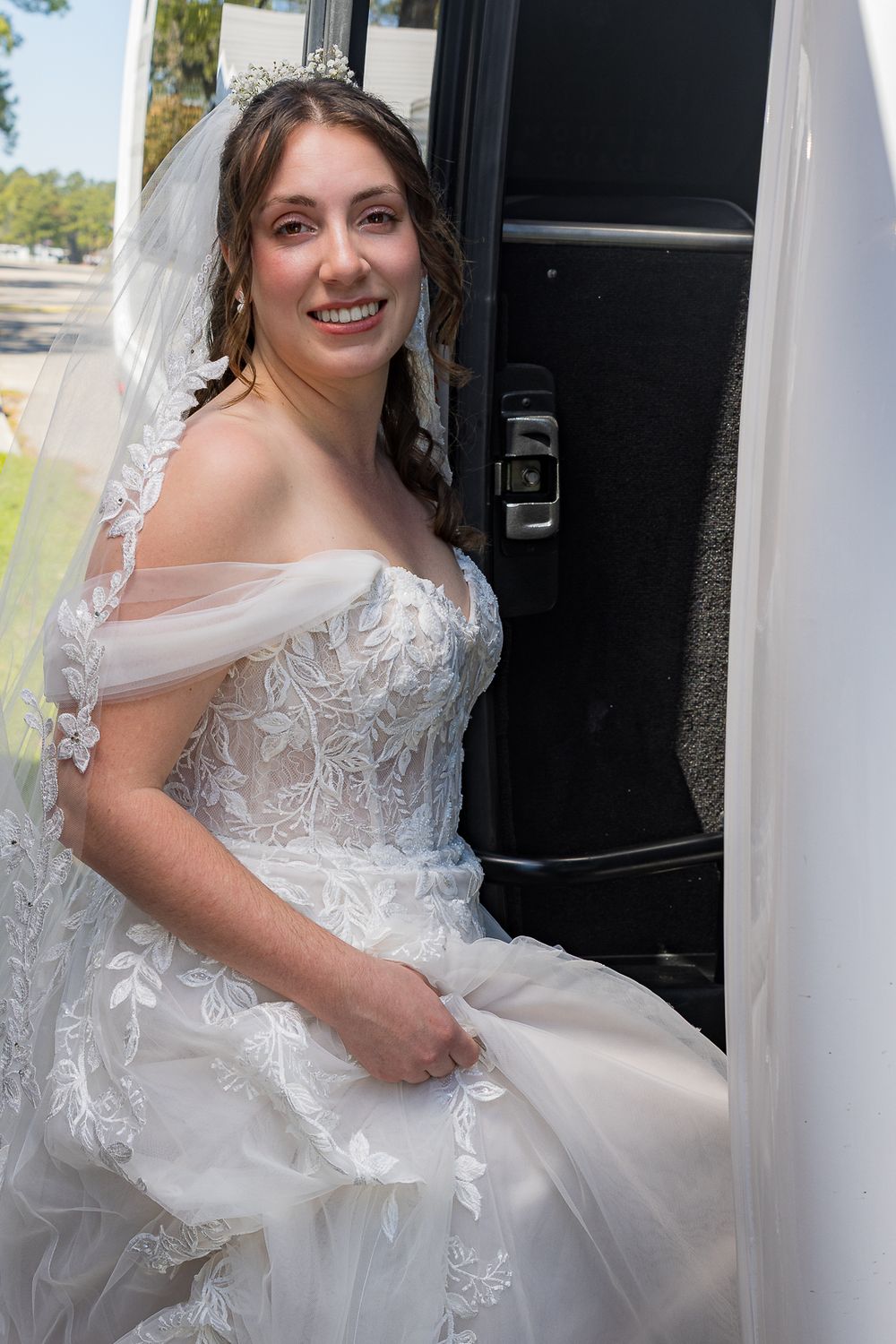 A bride in a white lace wedding gown with off-shoulder sleeves steps into a black vehicle on her special day.