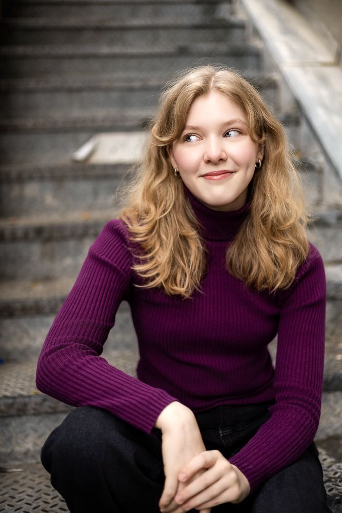 A person in a purple turtleneck sweater and dark pants sits on concrete steps while looking away with a gentle smile.
