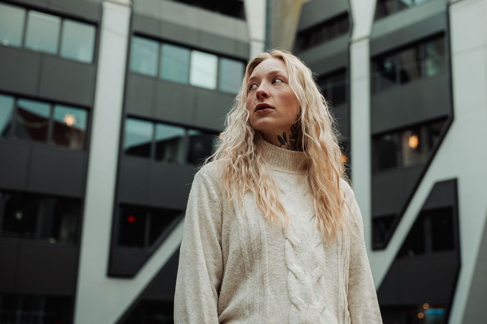 Person with long blonde wavy hair wearing a cream turtleneck sweater stands in front of modern architecture.
