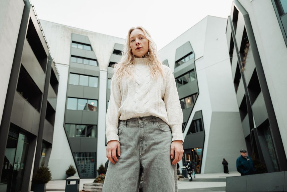 A person in a beige sweater and gray pants stands against a modern architectural backdrop with angular glass walls.