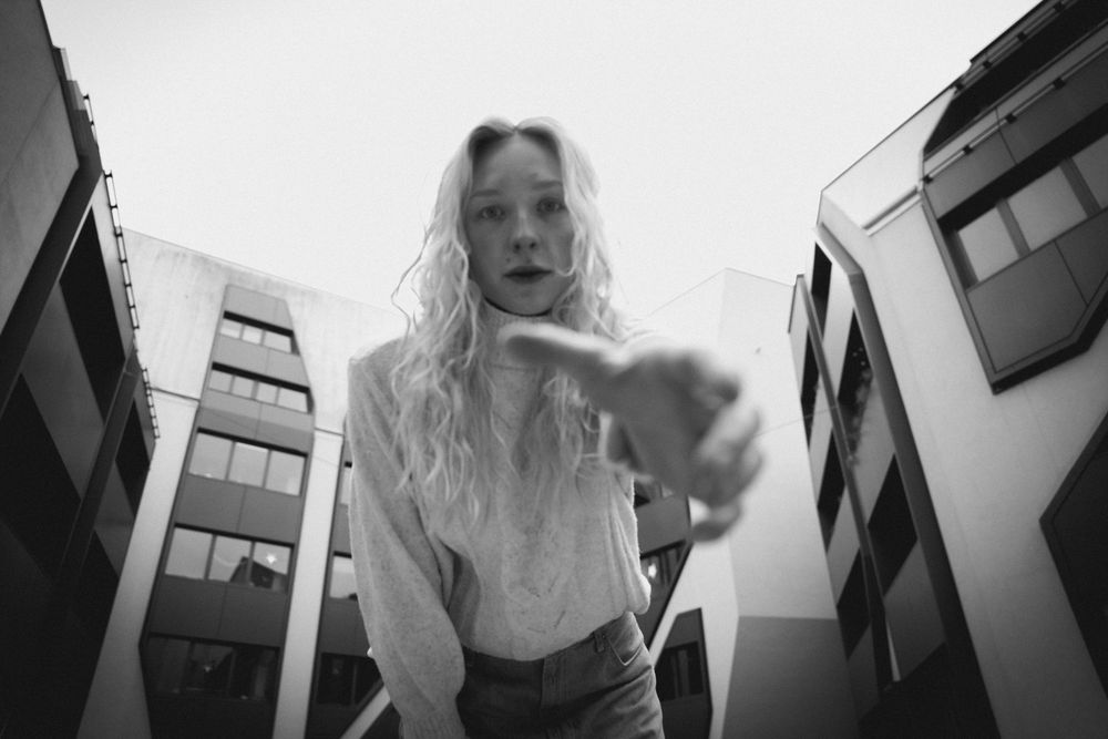 Black and white low angle photo of someone with long blonde hair pointing at camera between urban buildings.