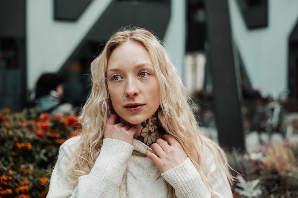 Person with wavy blonde hair wearing a white knit sweater poses outdoors near orange flowers and architectural elements.