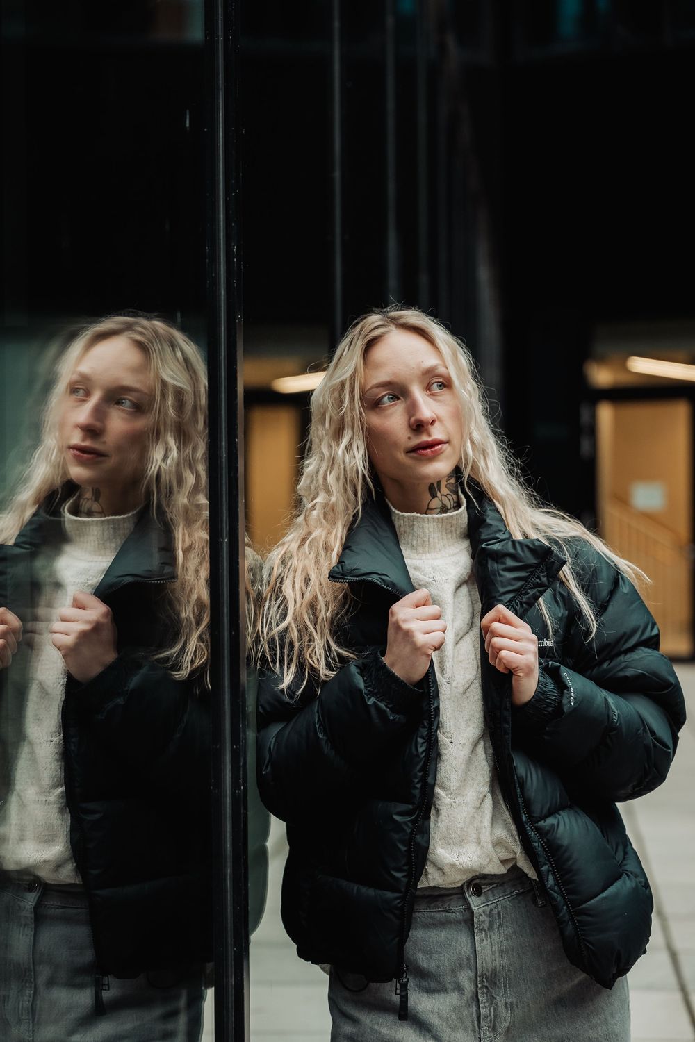 Person in a white sweater and black puffer jacket stands next to reflective glass surface in moody lighting.