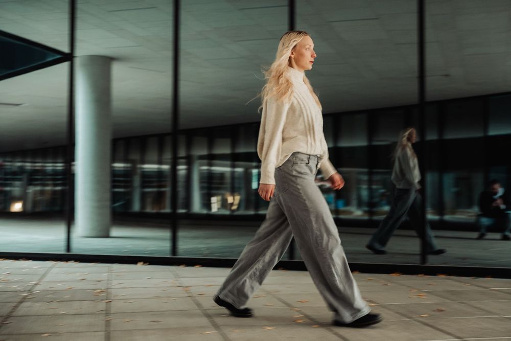 A person in casual business attire walks through a modern building with reflective glass walls and columns.