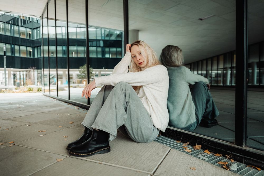 Two people sit back to back against a glass wall in an urban setting wearing casual clothes.