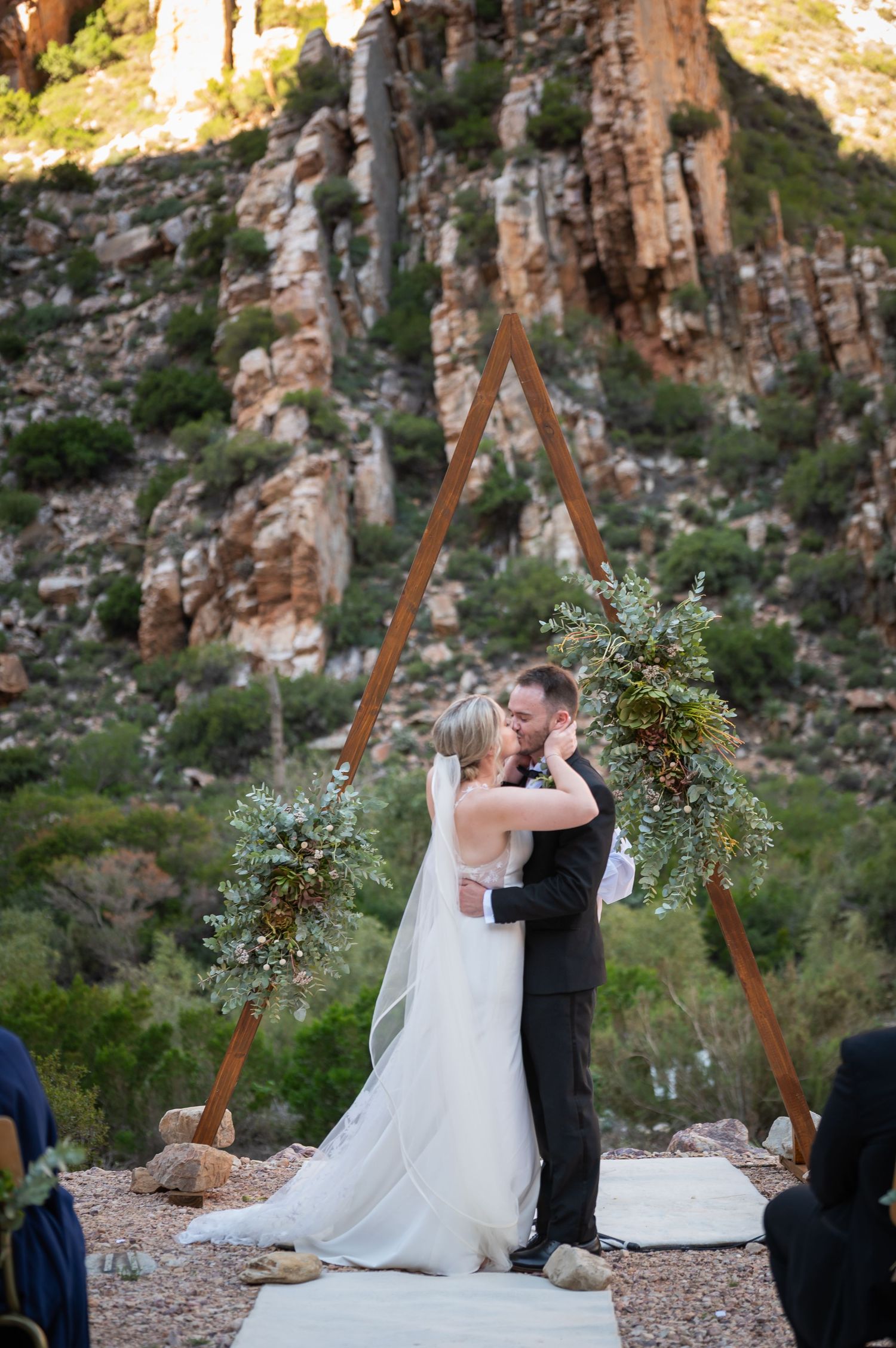 Bride and groom share first kiss under triangular wooden arch with greenery, set against stunning rocky mountain backdrop.