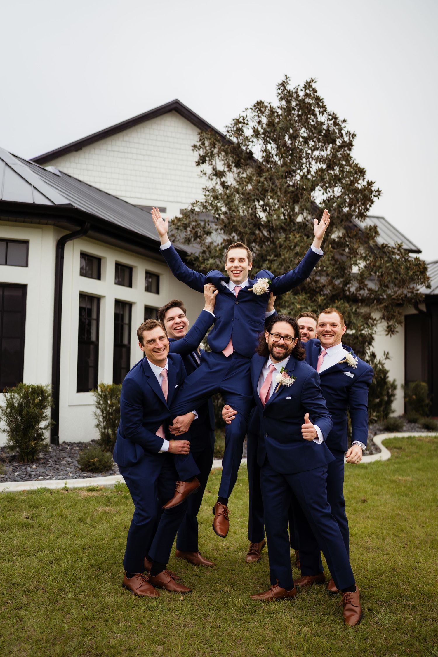 Groomsmen in navy suits pose playfully for a series of fun group photos on a lawn in front of a white building.
