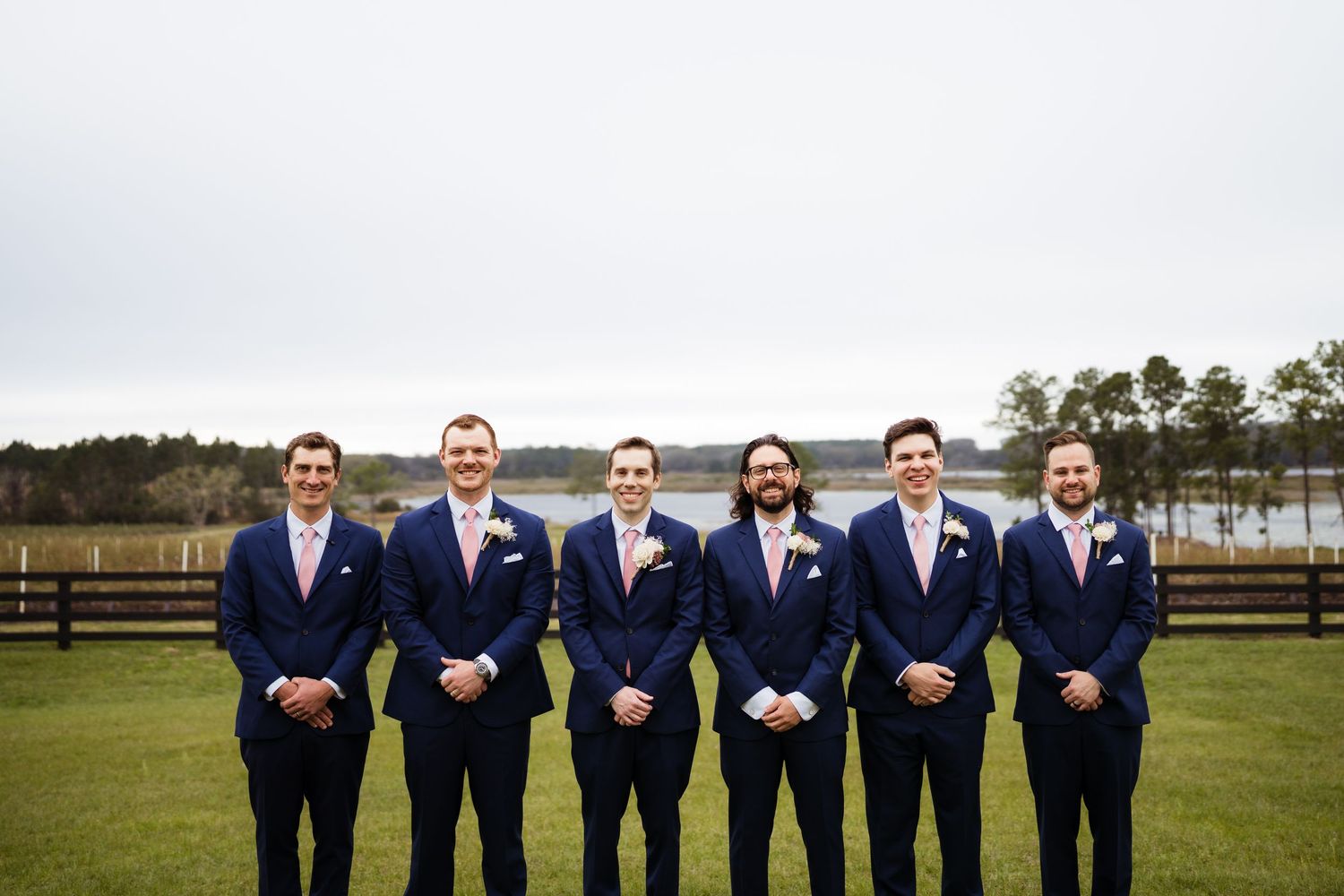 A group in navy blue suits poses together outdoors against a scenic rural landscape backdrop.