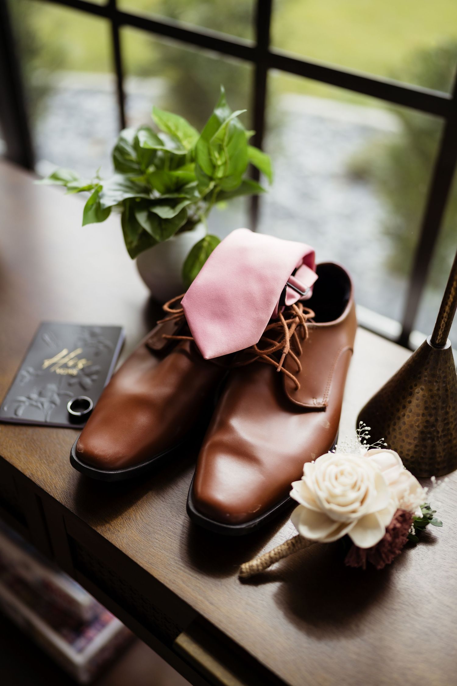 Brown leather dress shoes and white rose boutonniere arranged on dark wooden surface near window light.