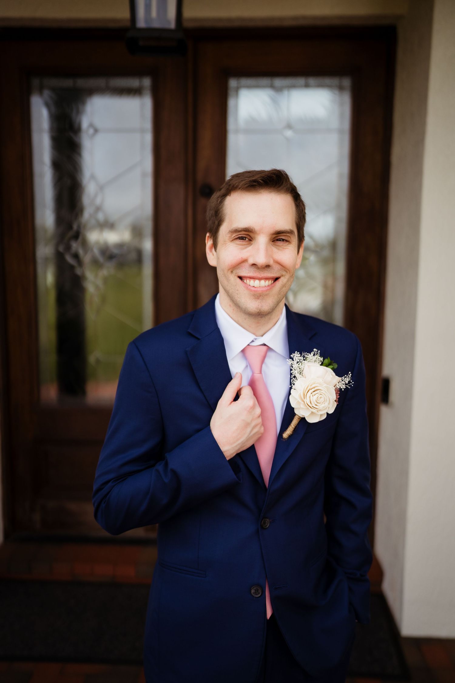 A groomsman in a navy suit with pink tie poses for wedding photos outside a building with white columns.