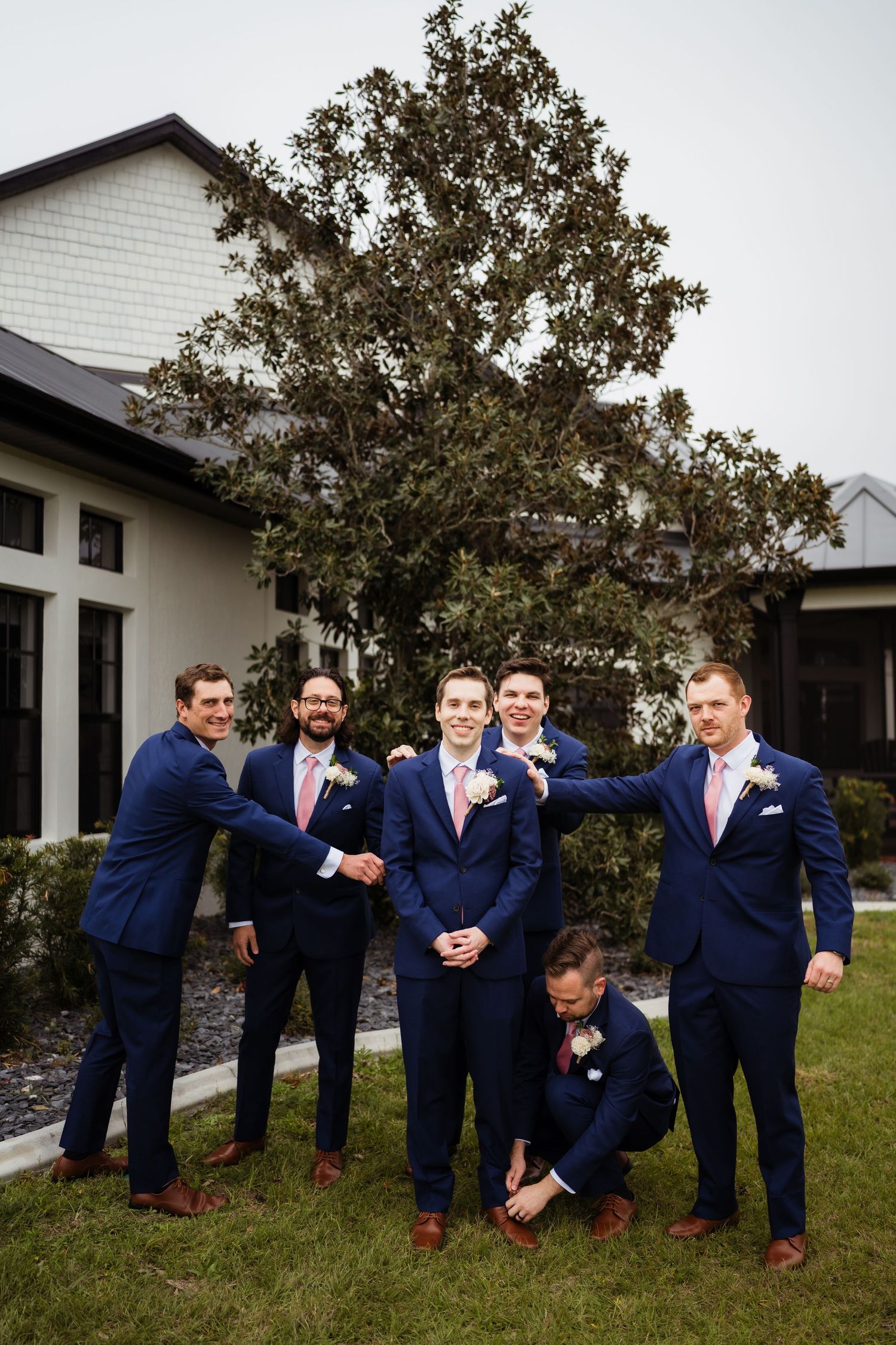 Groomsmen in navy blue suits pose playfully together in a series of candid shots outdoors by a white building.