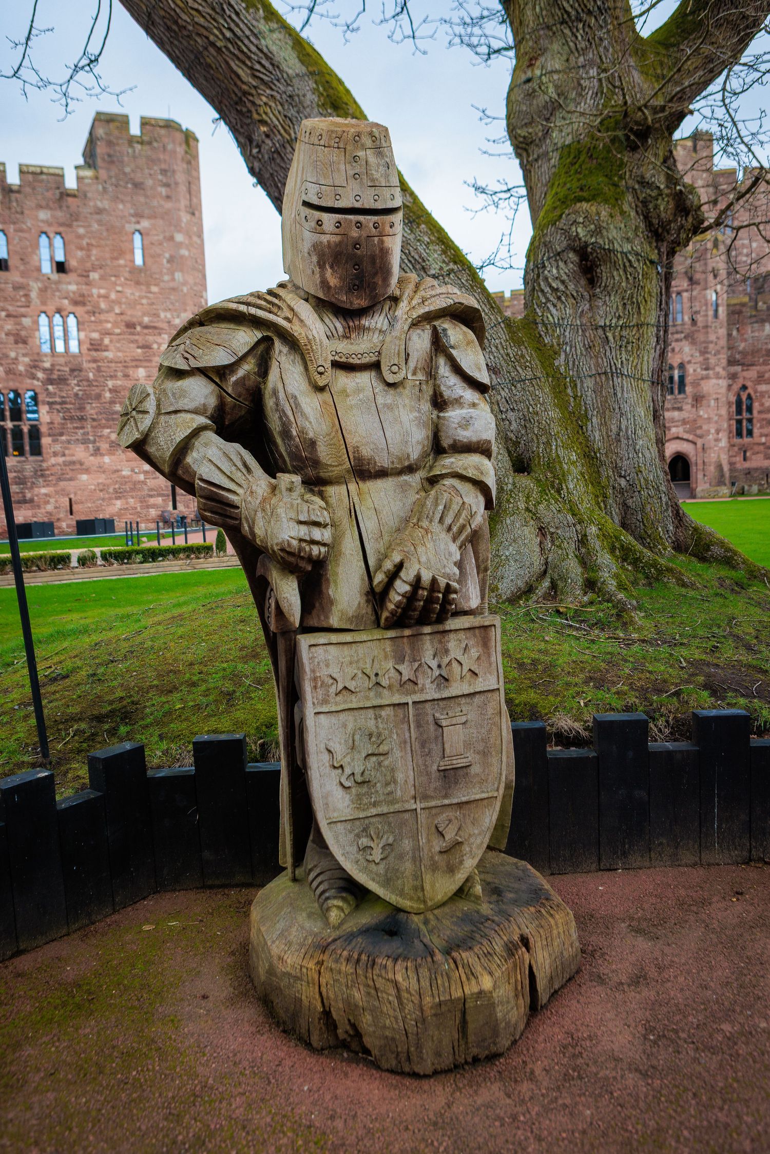 Carved wooden sculptures of medieval figures line a path near a historic red stone castle in the background.