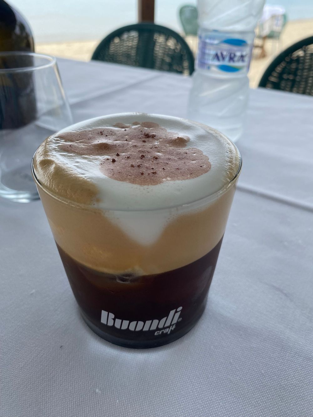 A layered coffee drink in a Buondi branded glass showing dark coffee topped with creamy foam and cocoa powder.