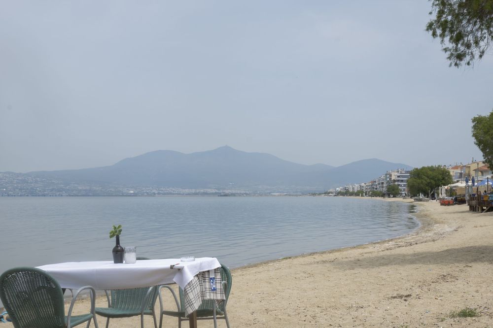 A tranquil beachfront scene with empty tables overlooking a calm bay and distant mountains in the background.